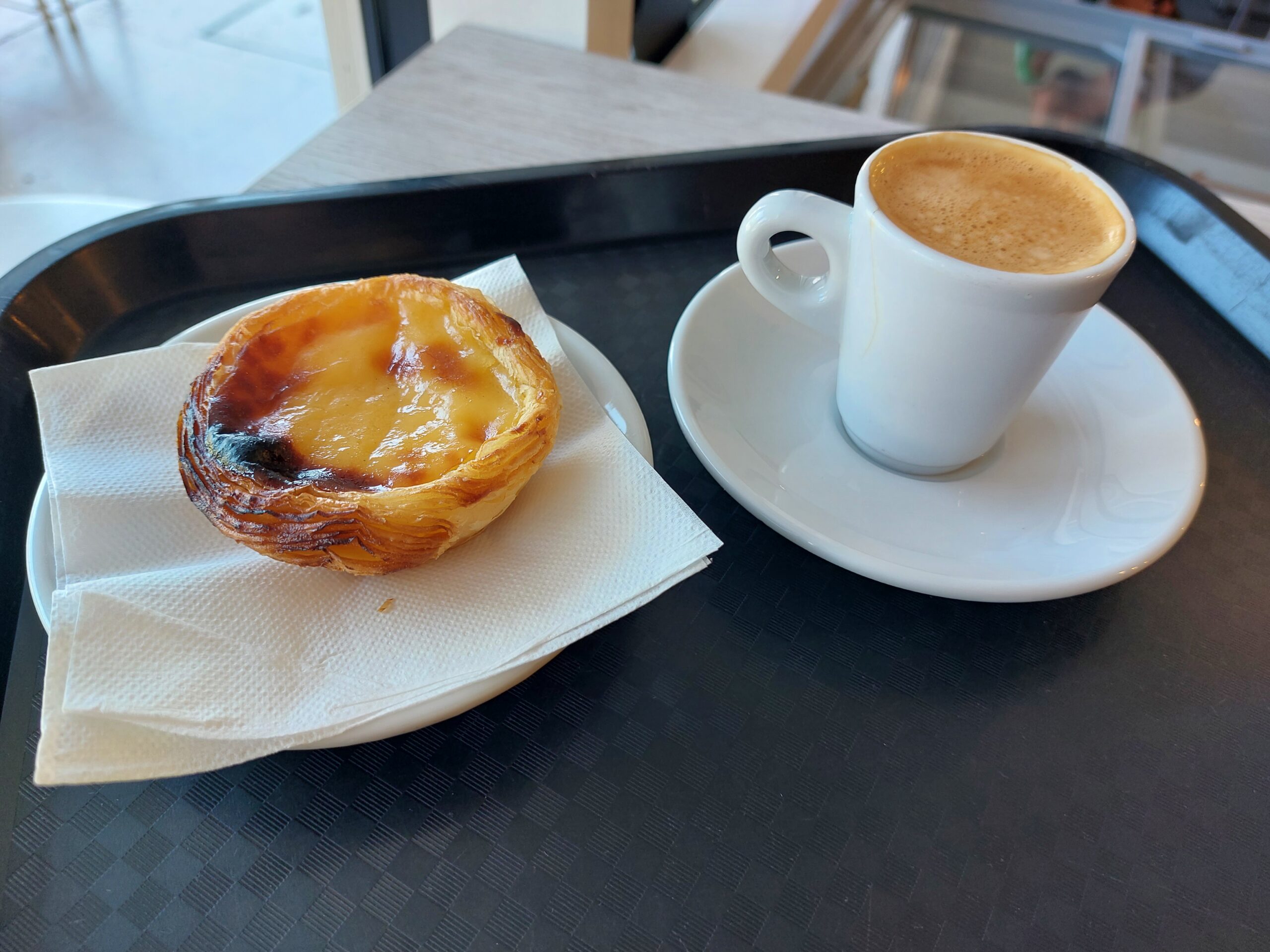 Pastel de nata on a paper napkin beside a small cup of coffee with milk foam at a Lisbon café