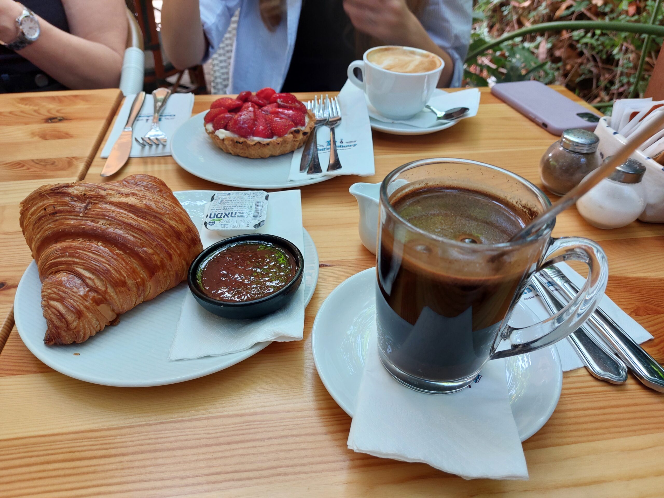 Black coffee in a glass mug with a croissant and jam, a strawberry tart and a cappuccino on a café table