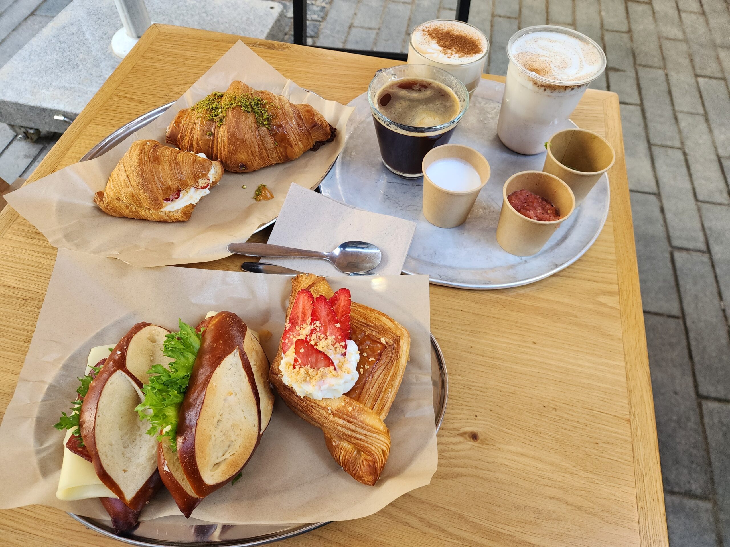 Café breakfast tray with two cinnamon-dusted lattes and a black coffee, next to croissants and a pretzel sandwich