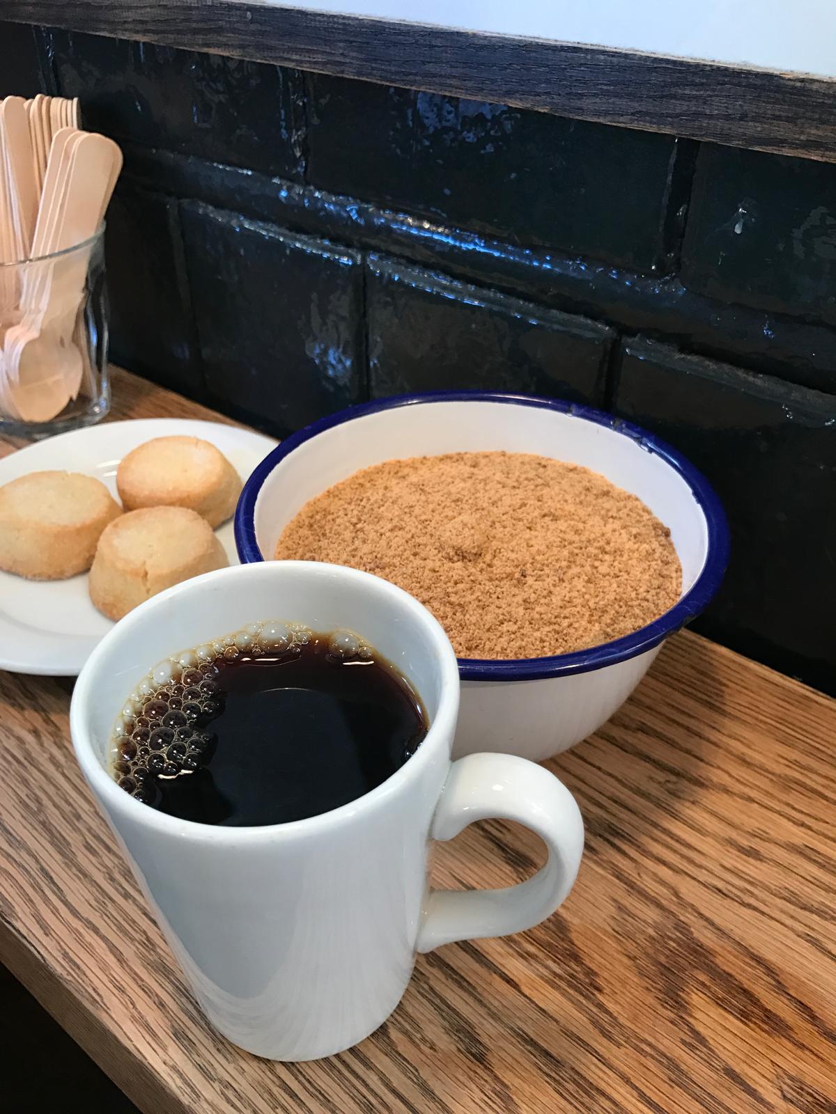 White mug of black filter coffee beside a bowl of brown sugar and shortbread biscuits on a wooden counter