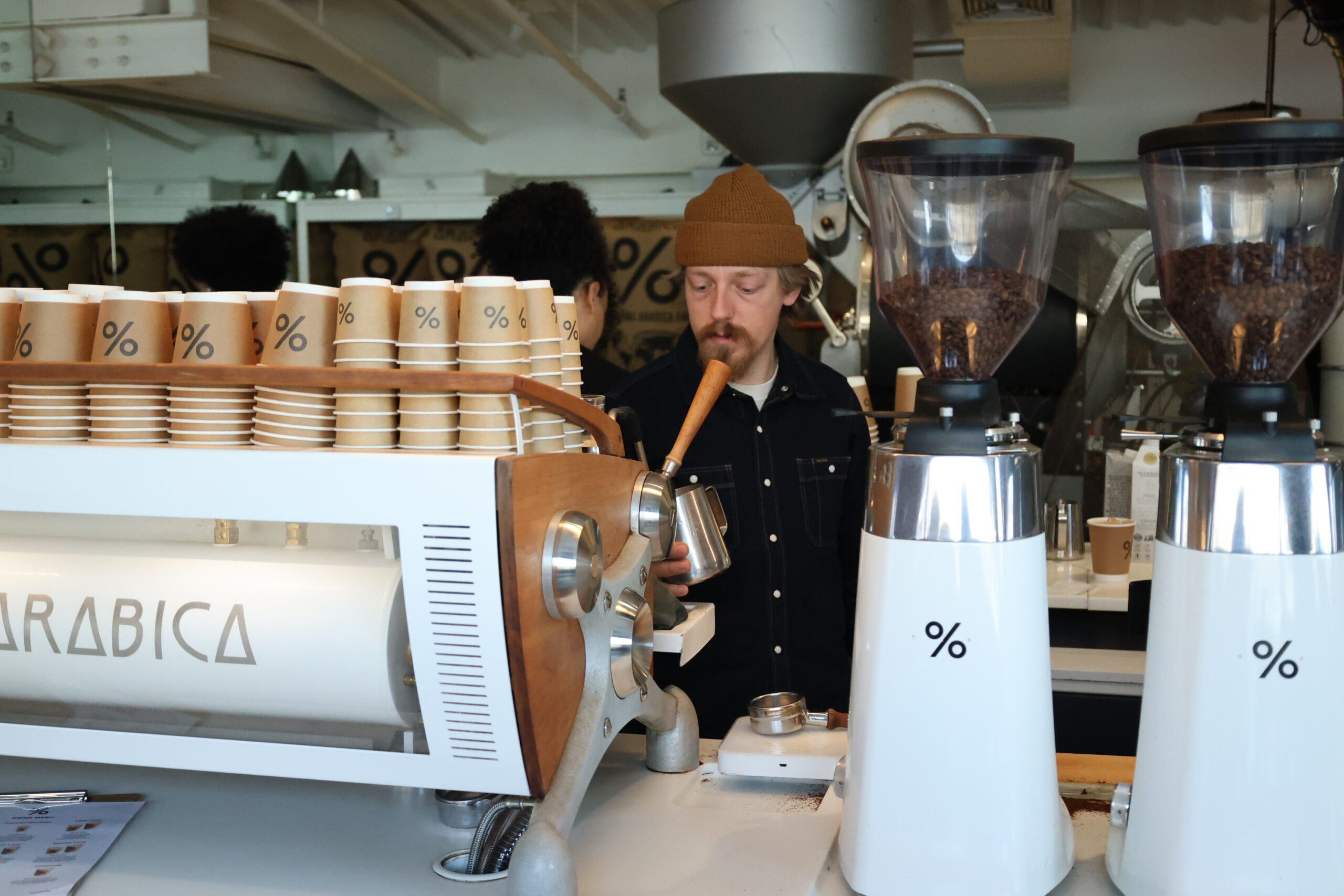 Barista steaming milk at an espresso machine beside two grinders with hoppers full of coffee beans