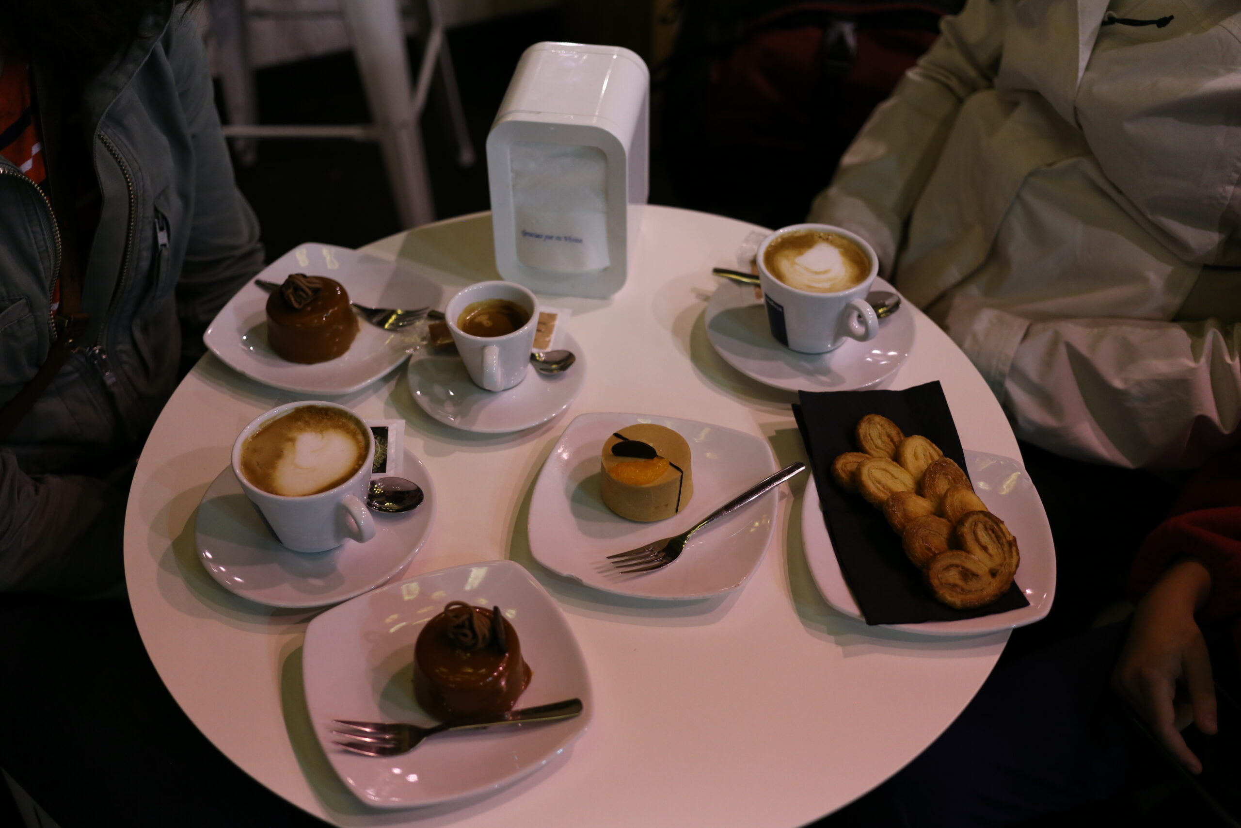 Two coffees with milk, a small black coffee, mousse cakes and palmier biscuits on a round café table at night