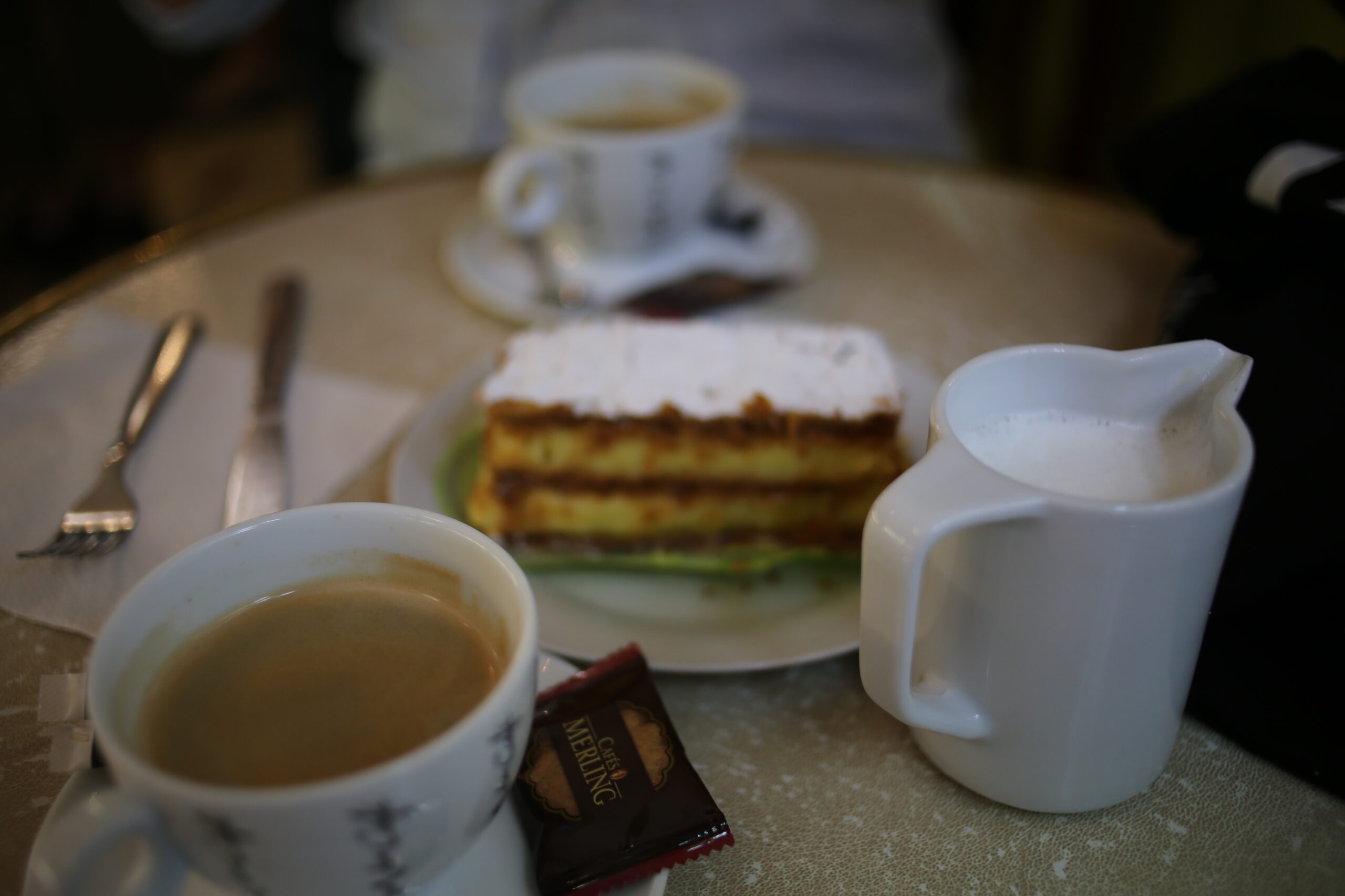 Cup of coffee with a wrapped biscuit, a jug of frothed milk and a slice of millefeuille on a café table