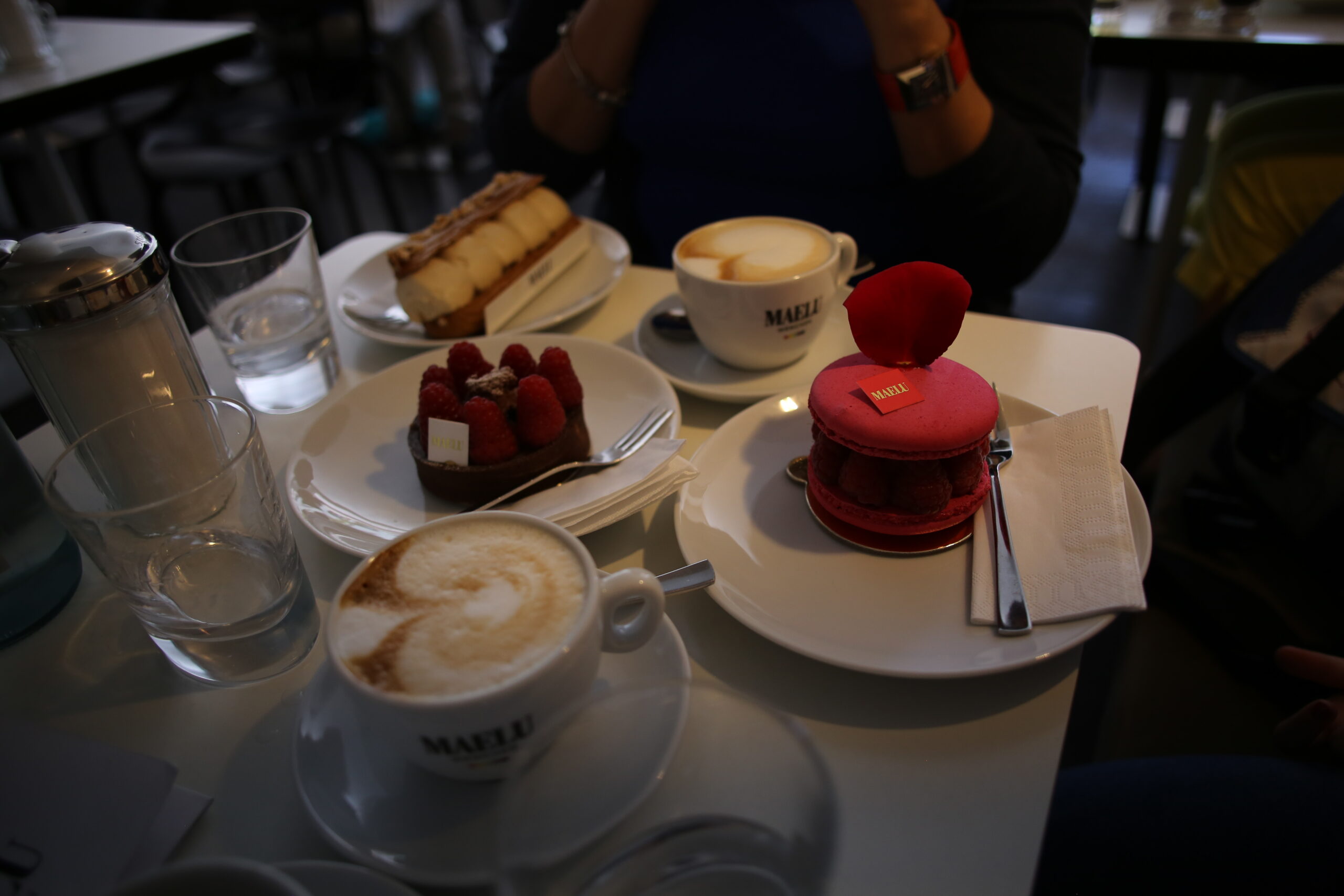 Two cappuccinos with latte art beside a raspberry tart and a pink macaron topped with a rose petal